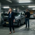 Woman on the phone in an office parking garage as her stalker ex waits beside her car holding a coffee cup