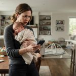 A mother holding her three-month-old baby while staring in shock at a phone her husband left on the table.