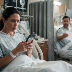 Woman awaiting cancer surgery looks at her phone while a neighboring hospital patient offers her a folded napkin.