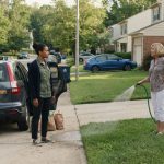 A woman carrying groceries from her car while a judgmental neighbor watches and accuses her of living off her tax money.