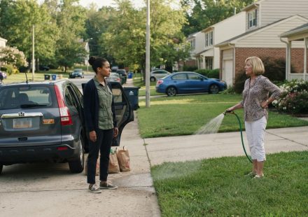 A woman carrying groceries from her car while a judgmental neighbor watches and accuses her of living off her tax money.