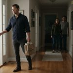 A husband carrying takeout enters his brother's house as his wife and brother emerge from the hallway.