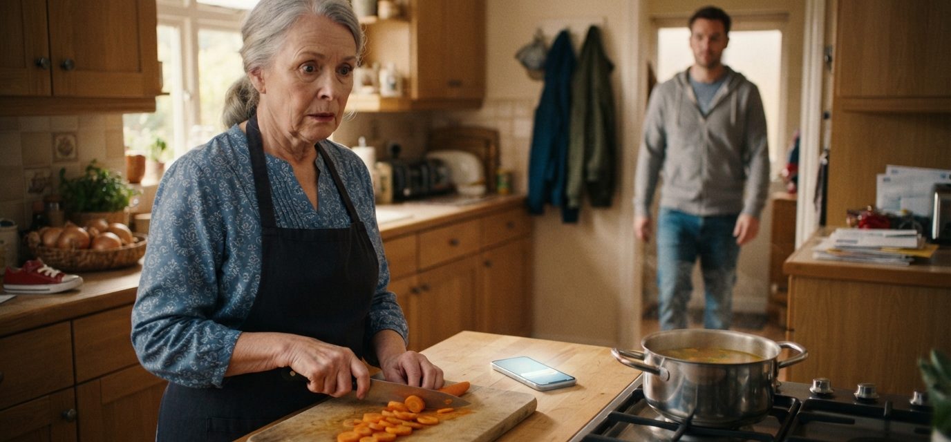 Older woman quietly staring at her son's illuminated phone while preparing soup in her kitchen as he approaches from the hallway.