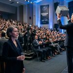 Inventor watching from the back of a corporate launch as an executive presents my $320,000 product concept onstage.
