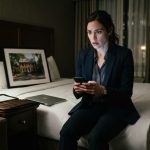 Professional woman staring at her phone in a Seattle hotel room after learning her family sold her Alexandria house without permission.