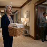 Grace holding a basket of folded laundry in the hallway as she overhears her son telling his wife that she lives off him.