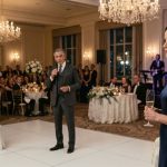 A composed woman beside a wedding cake watching the groom's wealthy father take the microphone during her younger sister's reception.