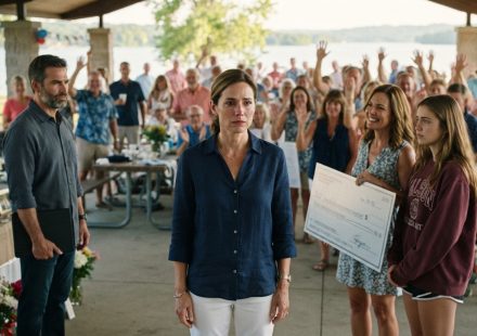 A woman standing apart from her family as her sister-in-law holds an oversized scholarship check during a lakeside family reunion.