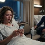 A woman in a hospital bed reading a troubling message on her phone while a male patient watches quietly from the neighboring bed.