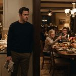A composed homeowner watches from his kitchen doorway as his family raises their glasses in celebration around his Thanksgiving dinner table.