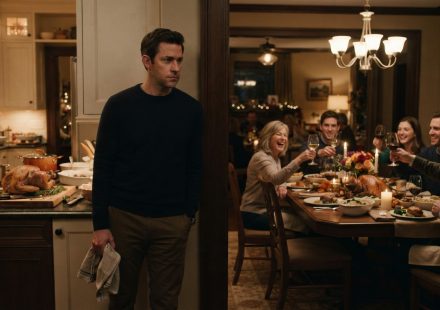 A composed homeowner watches from his kitchen doorway as his family raises their glasses in celebration around his Thanksgiving dinner table.