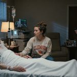 A daughter watches her dying father open his eyes in a hospice room while her wealthy brother stands nearby checking his phone.
