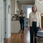 A 66-year-old mother stands beside a laundry basket in her daughter's hallway while her daughter, wearing workout clothes, confronts her impatiently.