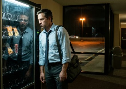 A tired business traveler stranded on a work trip with no car stands beside an almost empty hotel vending machine late at night.