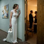 A bride in a white wedding dress stands barefoot in a church hallway holding her heels while secretly overhearing her fiancé speaking with his sister before their wedding.