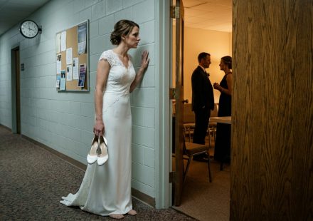 A bride in a white wedding dress stands barefoot in a church hallway holding her heels while secretly overhearing her fiancé speaking with his sister before their wedding.