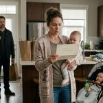 A mother holding one of her triplet babies while staring at an unexpected wedding invitation as her concerned husband stands nearby.