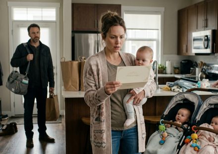 A mother holding one of her triplet babies while staring at an unexpected wedding invitation as her concerned husband stands nearby.