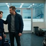 A worried father approaches his teenage son beside a hockey arena bathroom sink after receiving an urgent private message during practice.