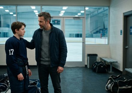 A worried father approaches his teenage son beside a hockey arena bathroom sink after receiving an urgent private message during practice.