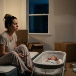A new mother sits beside her sleeping newborn in her newly purchased home, holding a phone after receiving an unexpected call from her mother.