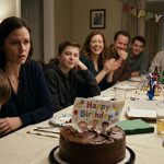 A shocked mother watches her nephew destroy her son's handmade birthday card in a chocolate cake while relatives laugh around the dinner table.