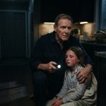 A worried retired grandfather answering his granddaughter’s emergency phone call late at night while rain falls outside.