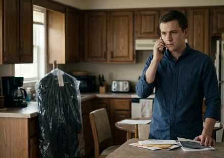 A 22-year-old college student standing in his kitchen, holding a phone with a hurt expression after his father announces he will miss his graduation.