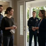 A worried mother holding a kitchen towel at her front door as a CPS investigator and police officer arrive, while her husband stands beside her and her mother-in-law watches from a parked car.
