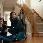 A frightened twelve-year-old boy holds his injured arm beside a staircase as his mother protects him and his aunt angrily points toward him.