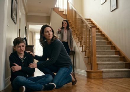 A frightened twelve-year-old boy holds his injured arm beside a staircase as his mother protects him and his aunt angrily points toward him.