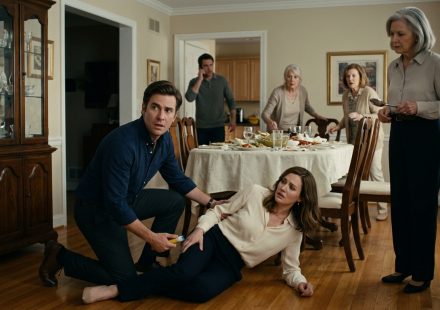 A frightened husband kneels beside his wife during a severe allergic reaction while his elderly mother stands holding a serving spoon at the family dinner table.