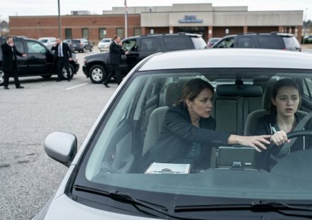 A frightened teenage girl grips the steering wheel during a driving test as her instructor reaches across to help her and black SUVs block the DMV parking lot.