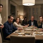 A distressed man sits at a family dinner table while his mother holds his hand and urges reconciliation, with his brother and other relatives seated nearby.