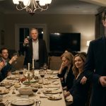A man holding a black USB drive stands beside a family reunion dinner table while his father raises a wine glass and relatives celebrate around him.