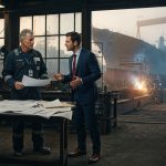 Veteran chief engineer facing a sharply dressed operations director inside a naval shipyard engineering office at dawn