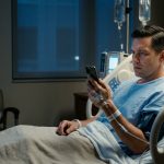 A man recovering from emergency appendix surgery lies in a hospital bed, holding a phone as his mother asks him to babysit his sister's children.