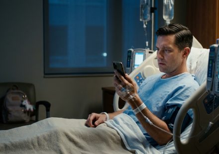 A man recovering from emergency appendix surgery lies in a hospital bed, holding a phone as his mother asks him to babysit his sister's children.