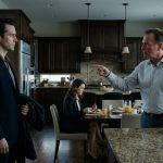 A composed man faces his angry father-in-law in an elegant kitchen while his wife sits silently at the breakfast table.