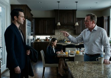 A composed man faces his angry father-in-law in an elegant kitchen while his wife sits silently at the breakfast table.