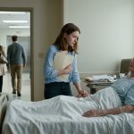 A seventeen-year-old girl standing alone in a hospital hallway holding her grandfather's medical chart while her family walks away toward the elevator.