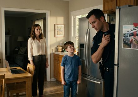 A concerned mother and her eight-year-old son watch as a police officer examines a fishing-trip photograph attached to their kitchen refrigerator.