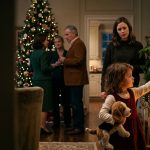 Mother protecting her young daughter in a decorated Christmas living room while her stern father watches nearby.