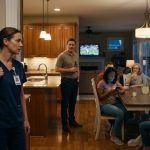 An exhausted nurse in medical scrubs arrives home to find six of her husband's relatives waiting around the dining table while her husband stands nearby.