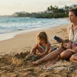 A mother sits on a Maui beach with her sleeping son and daughter, looking at her phone after her mom cancelled her Tahiti trip.