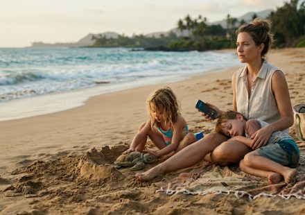 A mother sits on a Maui beach with her sleeping son and daughter, looking at her phone after her mom cancelled her Tahiti trip.