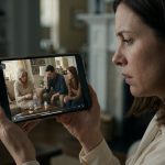 A shocked woman checking nanny-camera footage on her smartphone and discovering her mother-in-law handling a supplement bottle in the family living room.