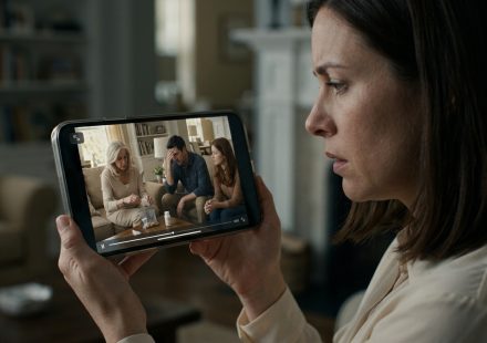 A shocked woman checking nanny-camera footage on her smartphone and discovering her mother-in-law handling a supplement bottle in the family living room.