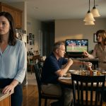 A disappointed young woman stands in her family kitchen while her parents ignore her career announcement and focus on her brother.