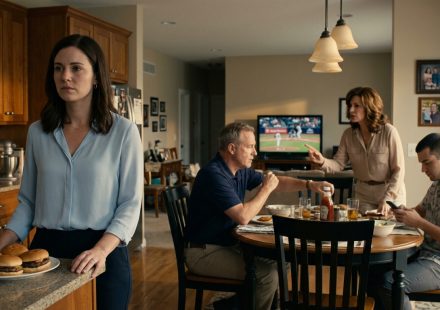 A disappointed young woman stands in her family kitchen while her parents ignore her career announcement and focus on her brother.